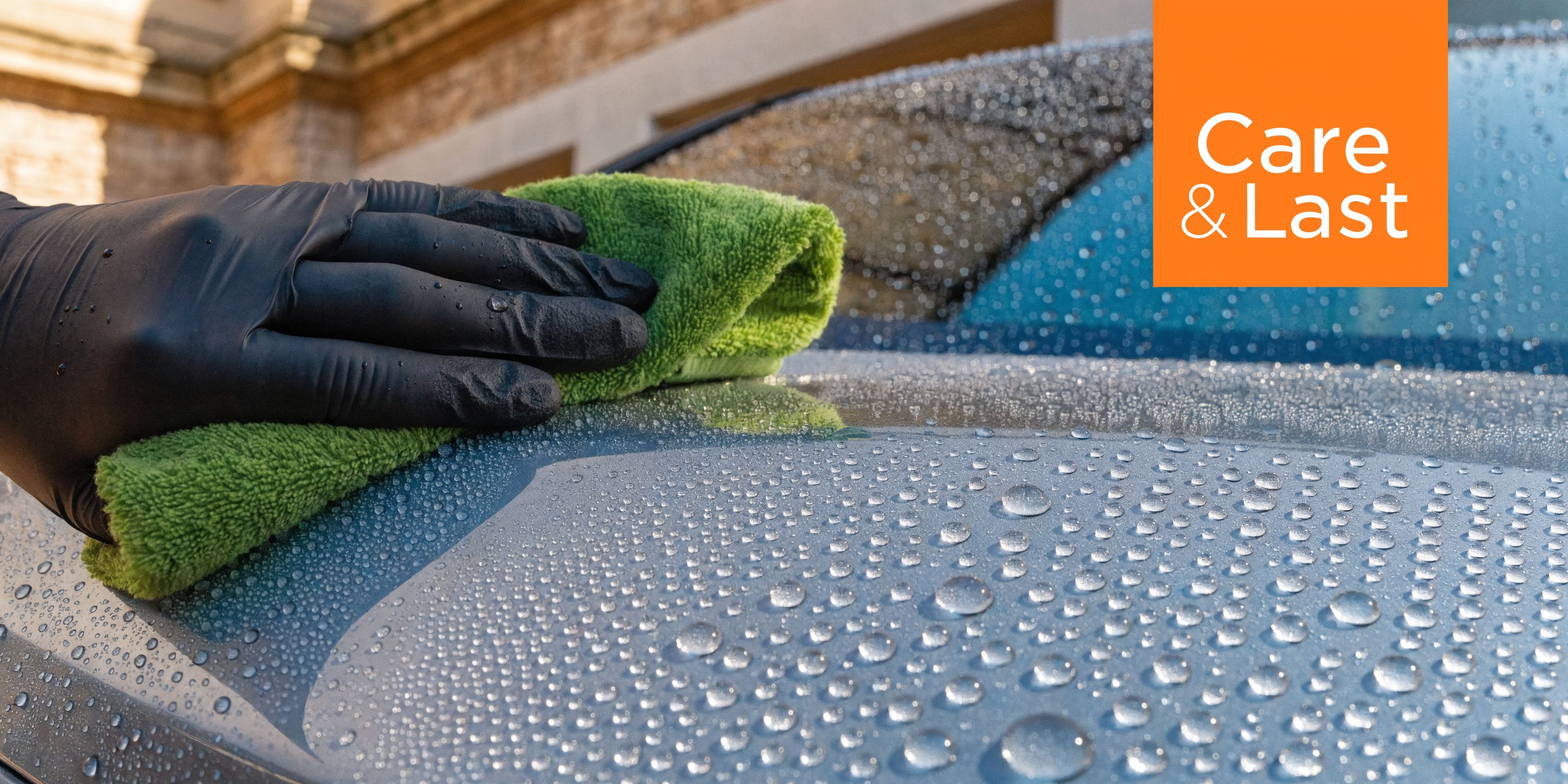 A hand wearing a black protective glove wipes a wet blue car hood with a green microfiber cloth.