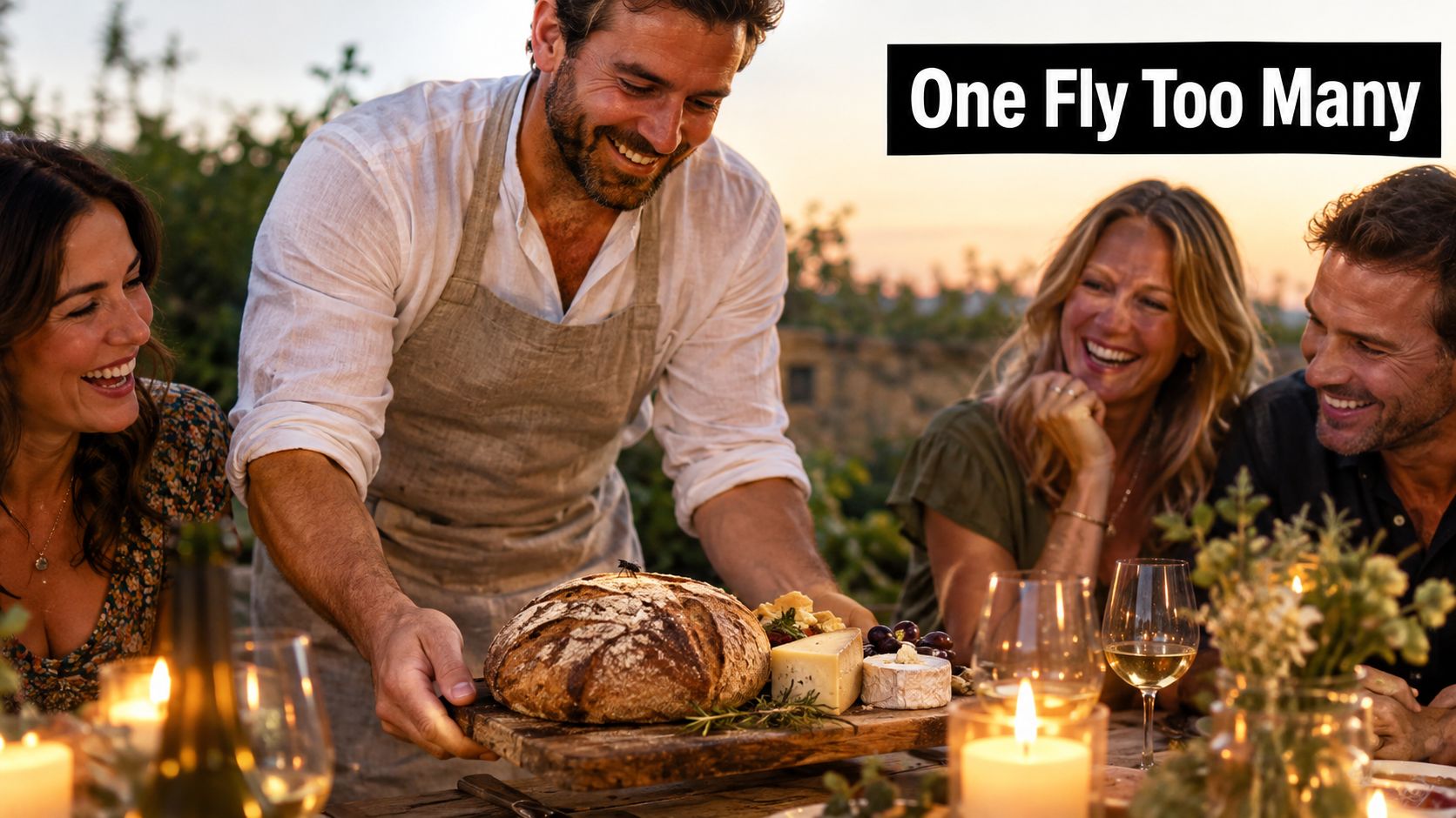 A man places a loaf of bread on a table where friends are gathered to dine outdoors.