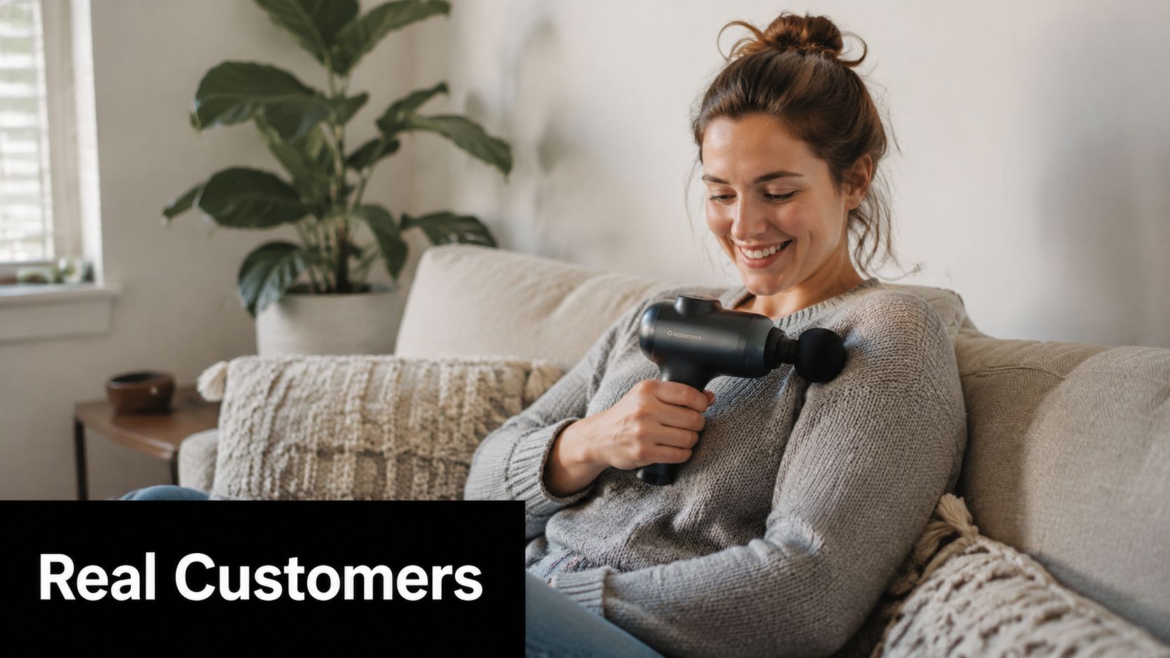 A happy woman sitting on a beige couch using a black massage gun on her shoulder
