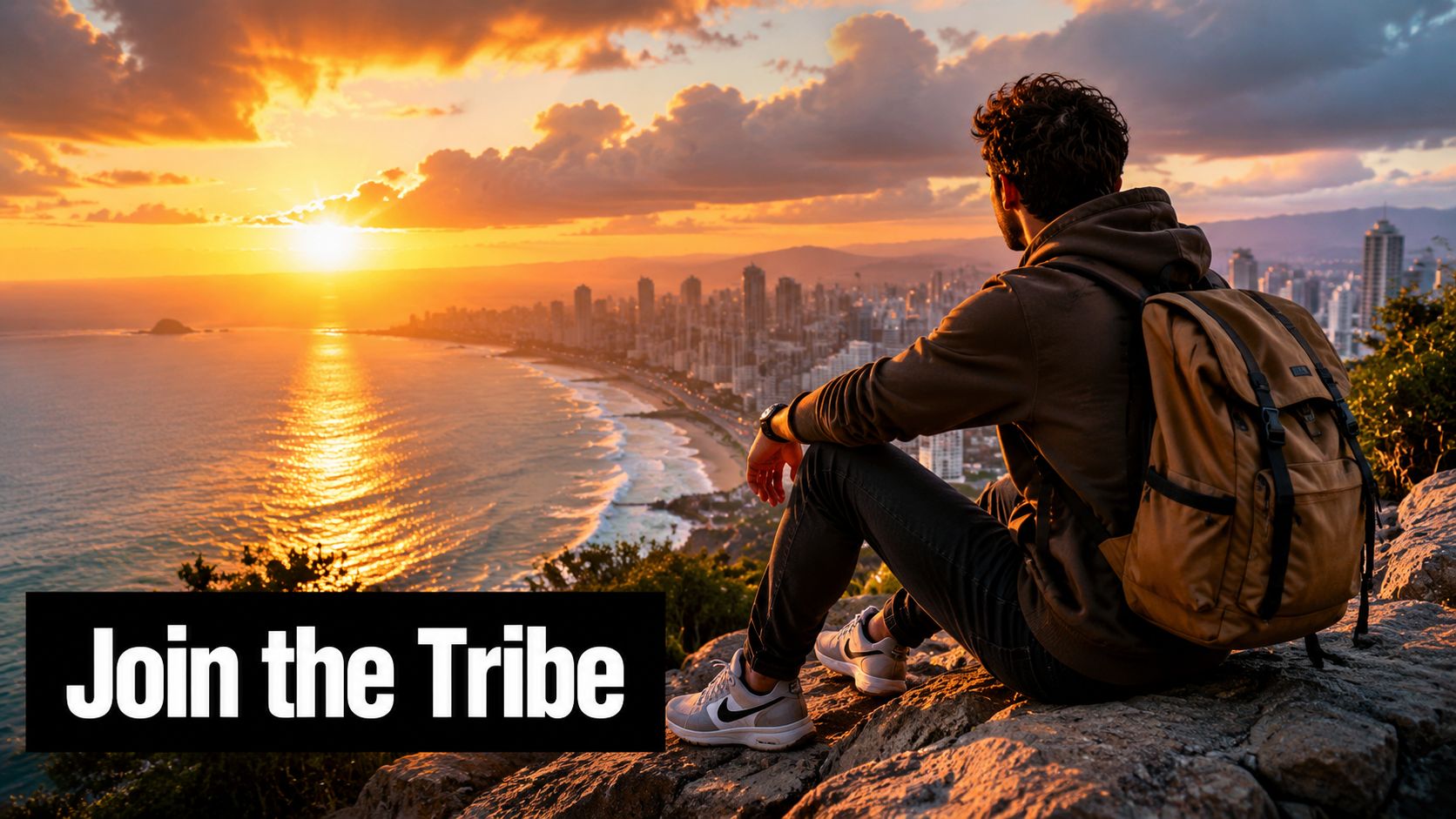 A man with a backpack sits on a rocky overlook watching the sun set over a city skyline.