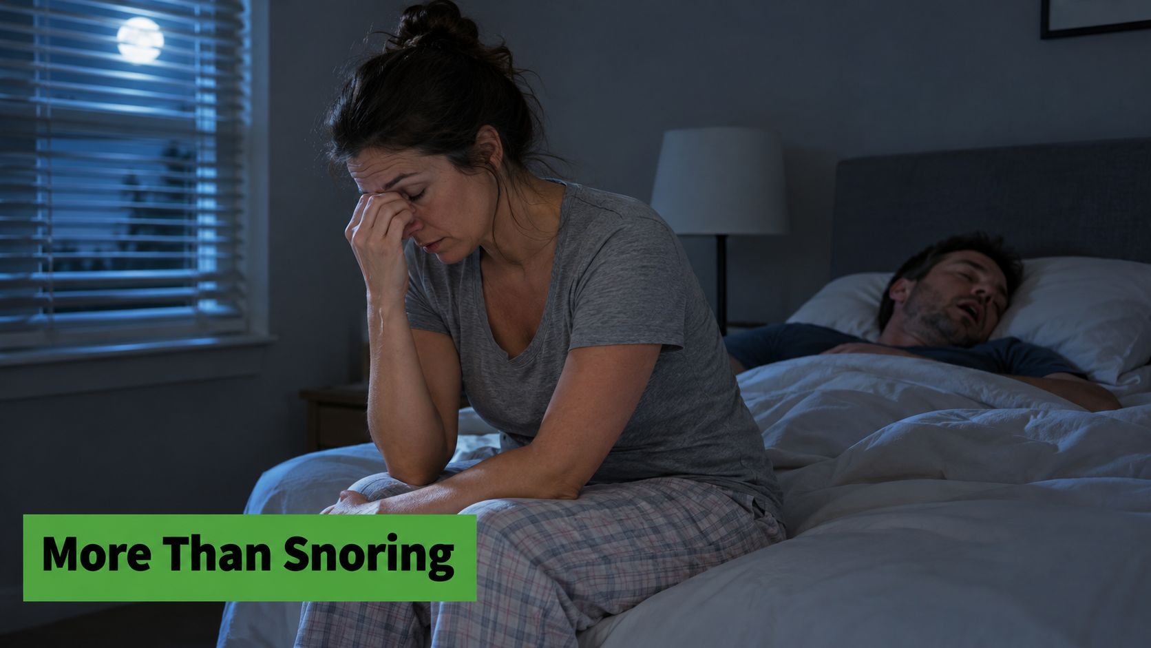 An exhausted woman sits on her bed looking distressed while her partner snores in the background.