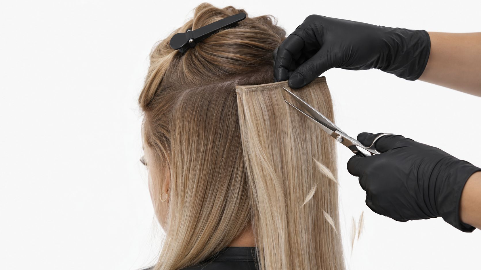 A professional hair stylist in black gloves trims long blonde hair extensions on a woman's head.