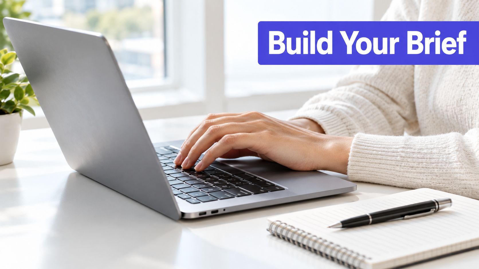 A person wearing a white sweater typing on a silver laptop at a desk with a notebook.
