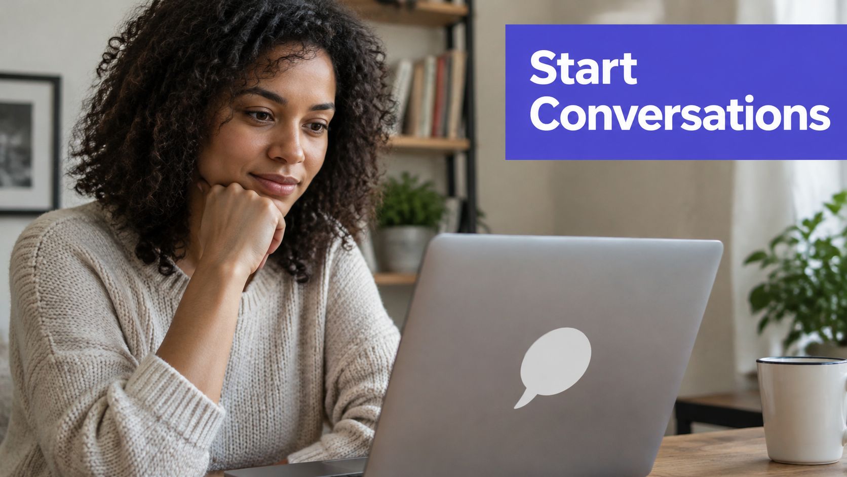 A woman with curly hair sits at a desk looking at a laptop with a speech bubble sticker.