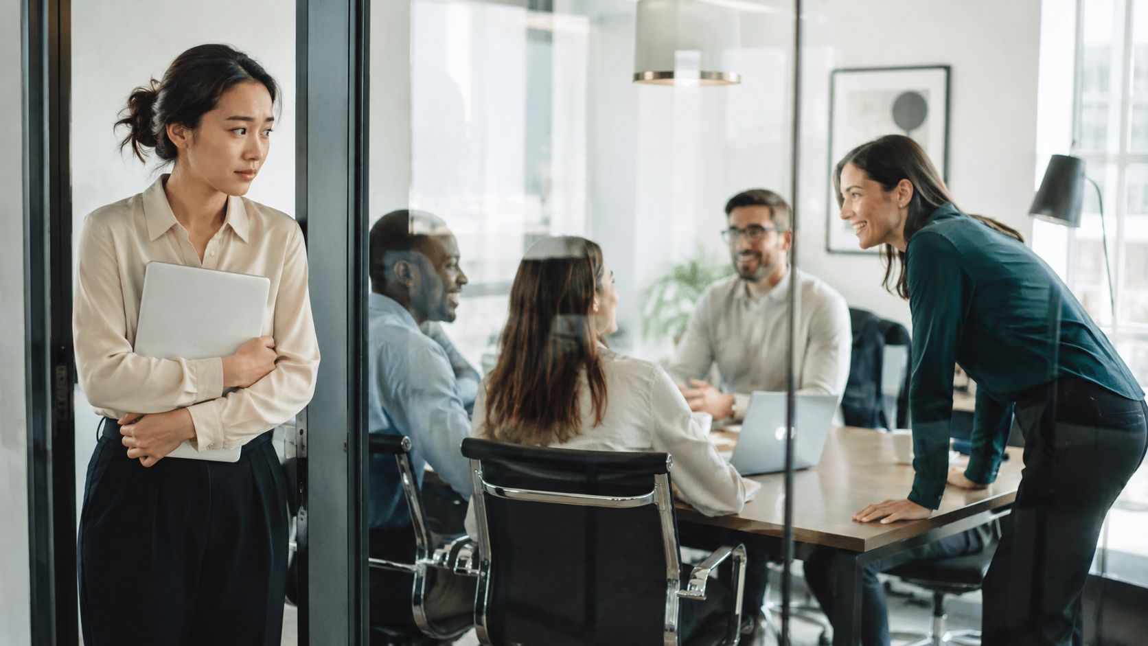 A lonely woman standing outside a glass office partition while colleagues collaborate during a business meeting.