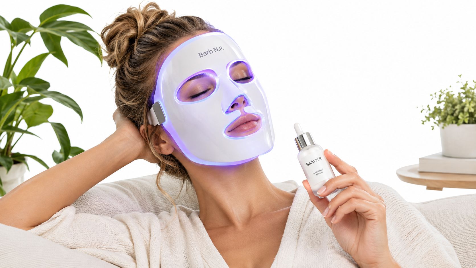 A woman wearing a glowing LED light therapy face mask while holding a bottle of facial serum.