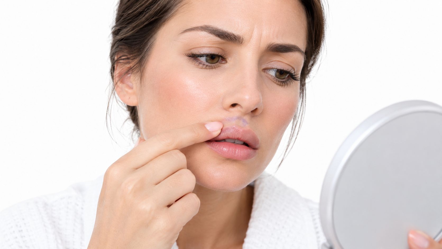 A concerned woman examining her lip filler migration while looking in a small round mirror at home.