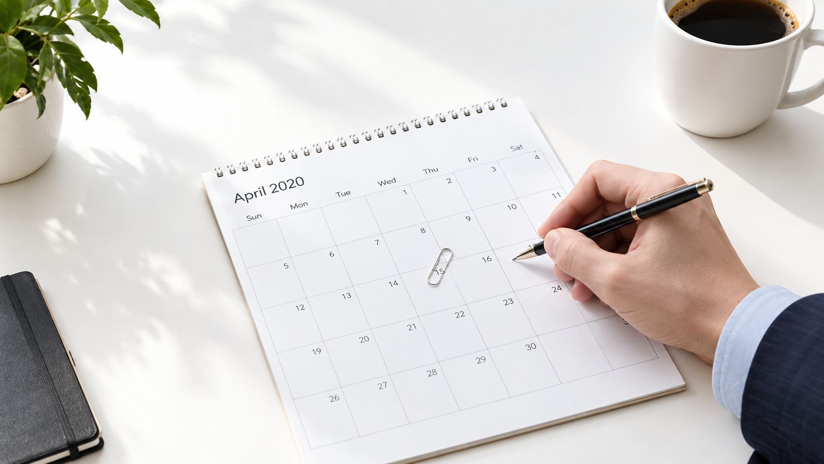 A businessman writing on an April 2020 calendar desk planner with a black pen and coffee nearby.