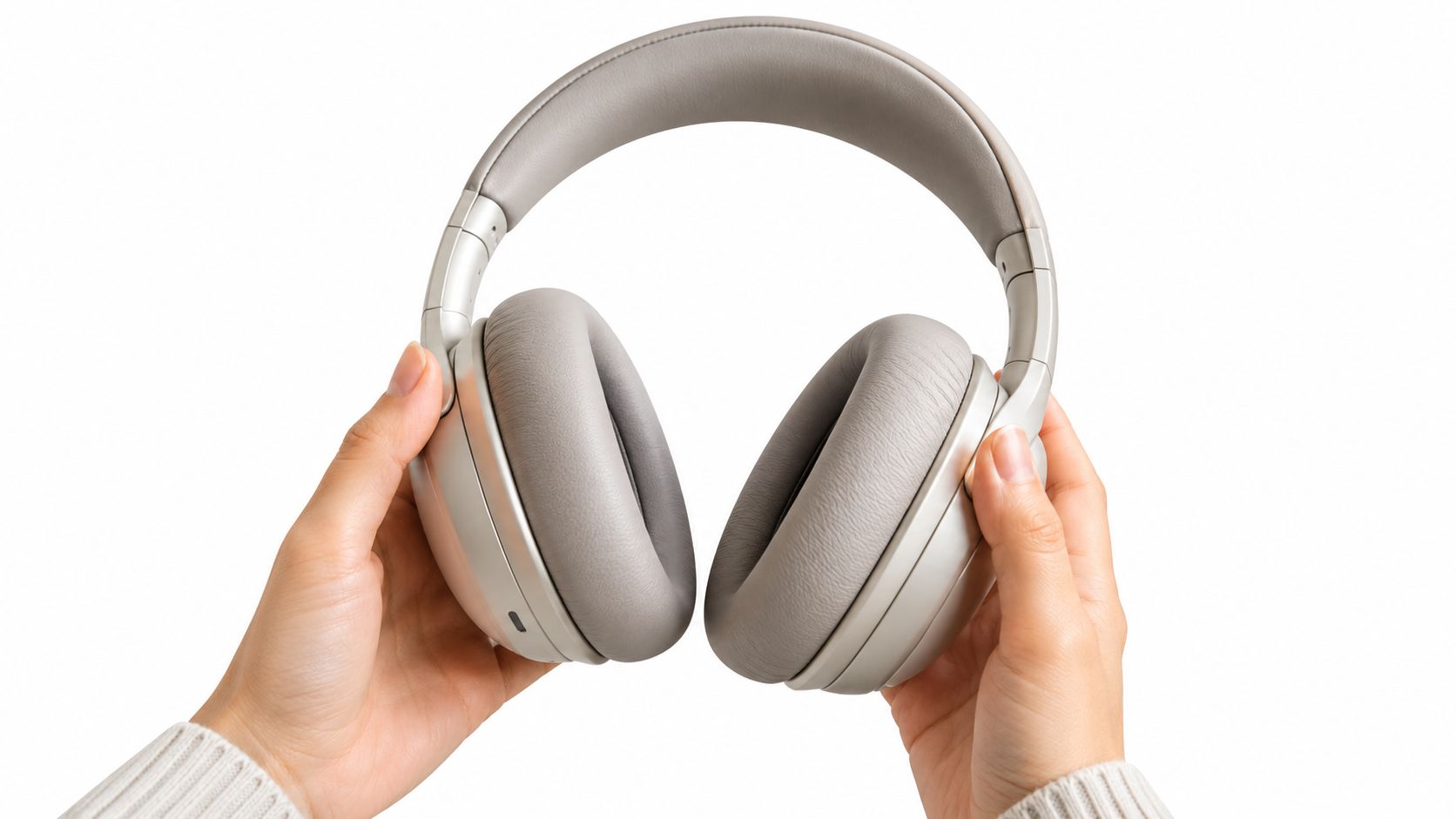 A person holding a pair of light gray wireless closed-back headphones against a plain white background.