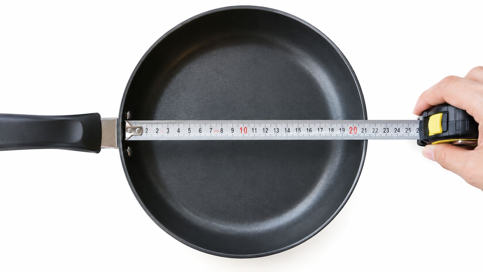 A hand using a metal measuring tape to determine the diameter of a black frying pan.