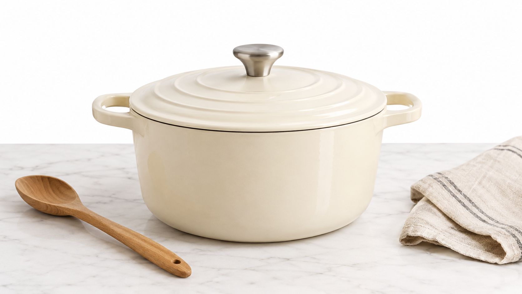 A beige enameled cast iron Dutch oven with a wooden spoon and a linen towel on marble.