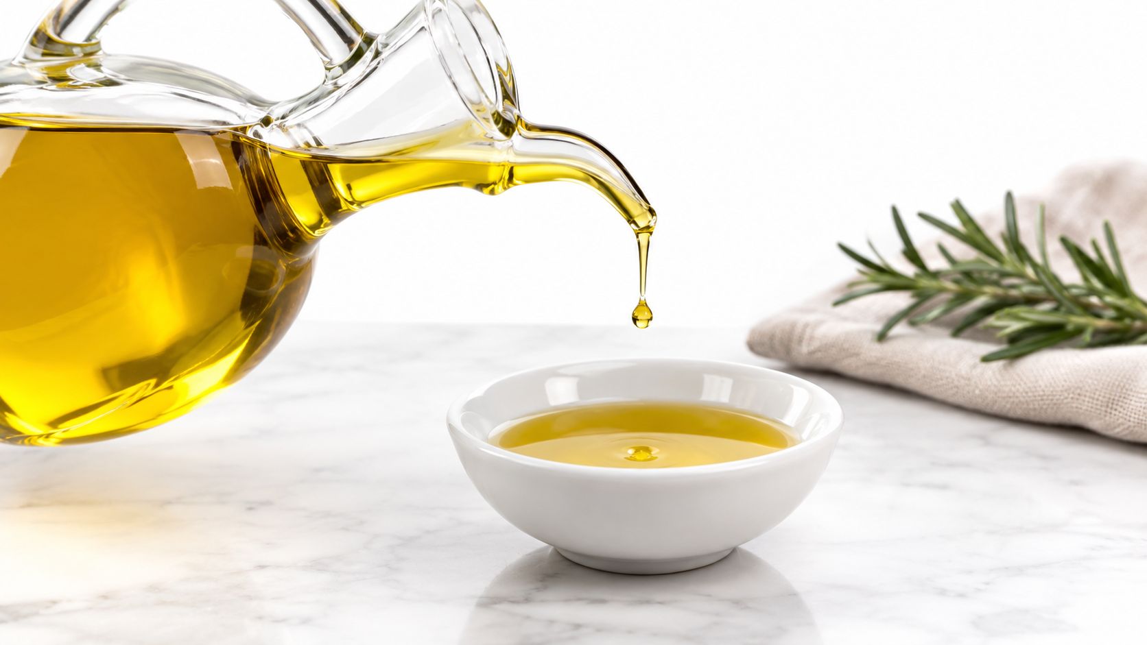 A glass cruet pouring golden olive oil into a small white bowl on a marble surface.