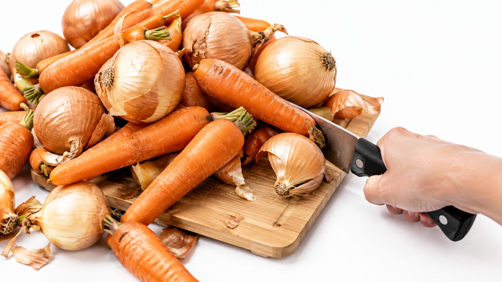 A hand holding a knife preparing to chop fresh orange carrots and brown onions on a board.