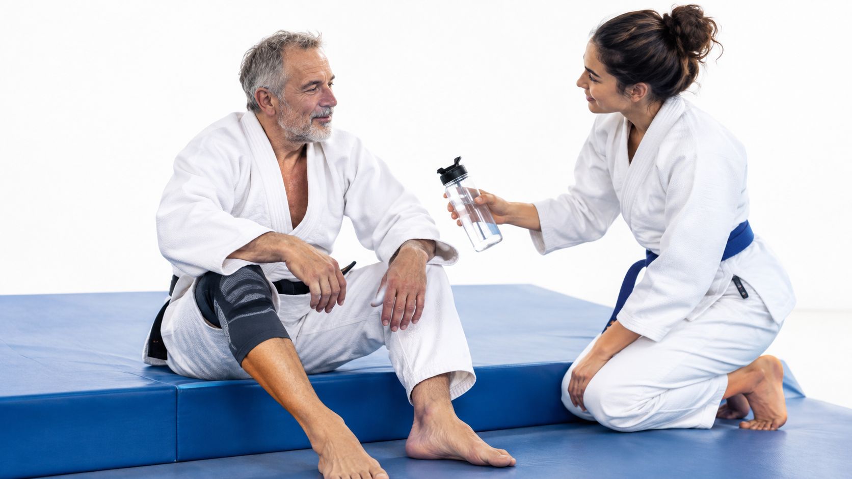 A female martial arts student offers a water bottle to an older man resting on a blue mat.