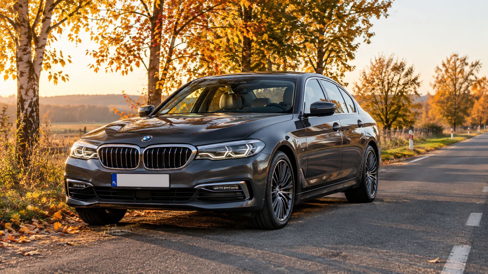 A sleek dark grey BMW luxury sedan parked on the side of a road during a beautiful autumn sunset.