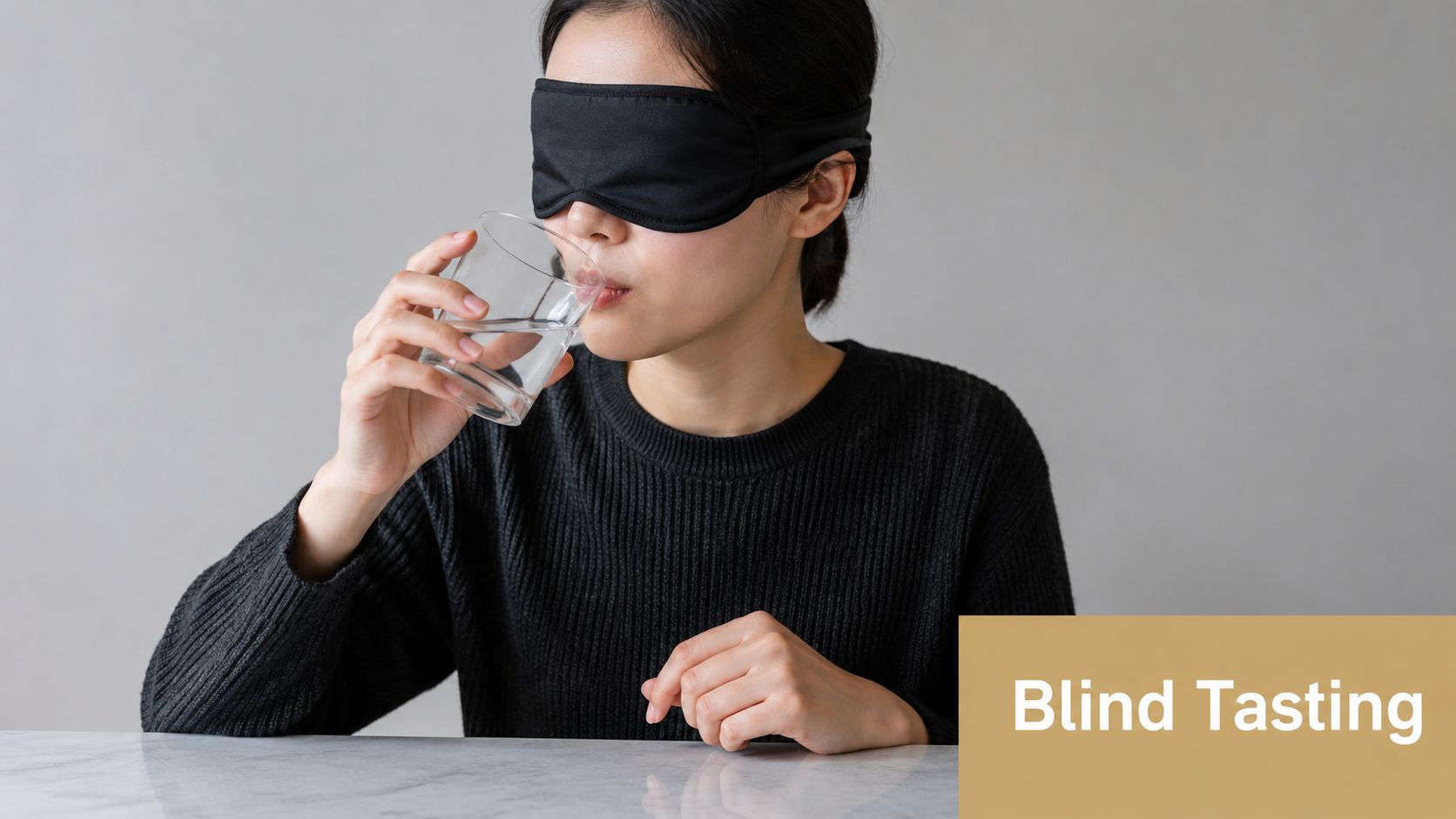 A woman wearing a black sleep mask is drinking water from a clear glass during a blind tasting.
