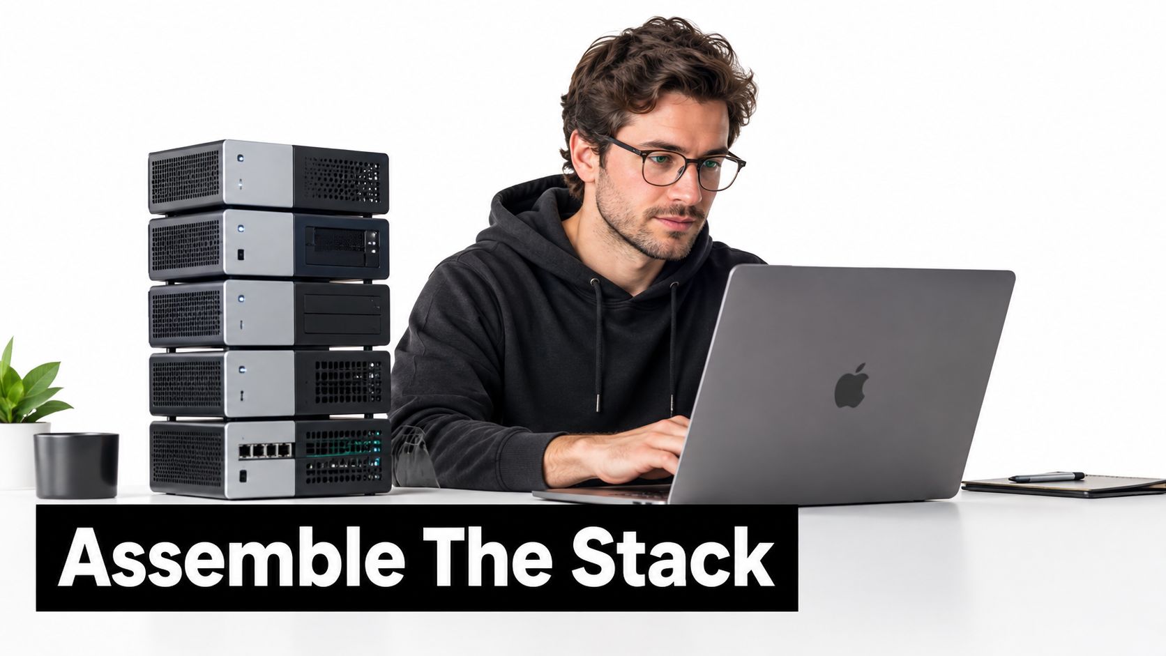 A professional developer sitting at a desk with a laptop next to a vertical stack of servers.