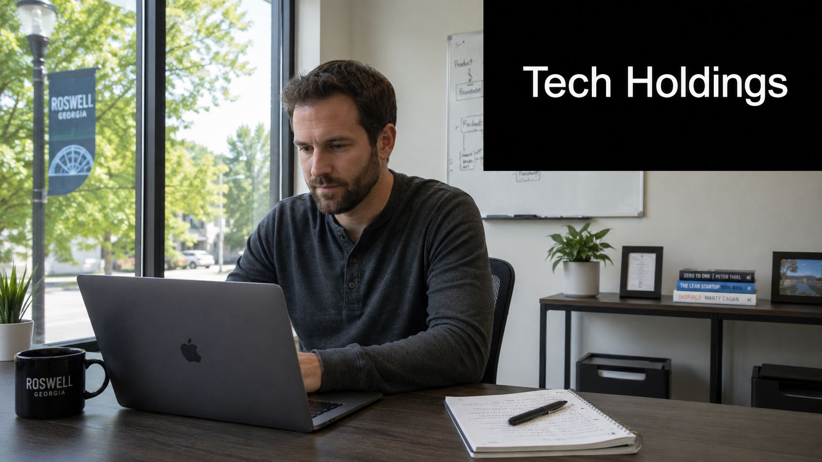 A man with a beard sits at a desk working on his laptop in a bright office.