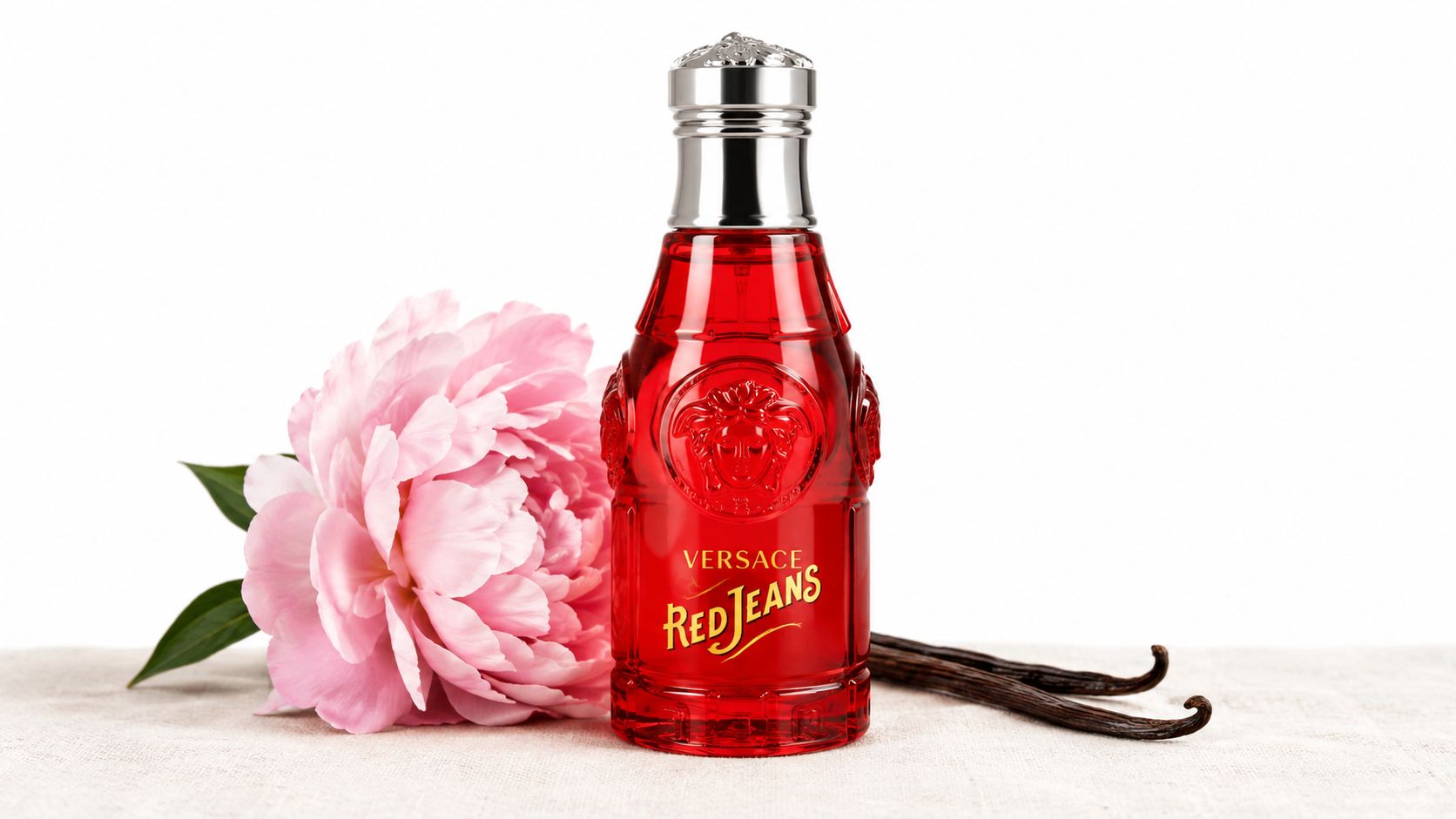 A bottle of Versace Red Jeans perfume placed next to a pink peony and vanilla bean pods.