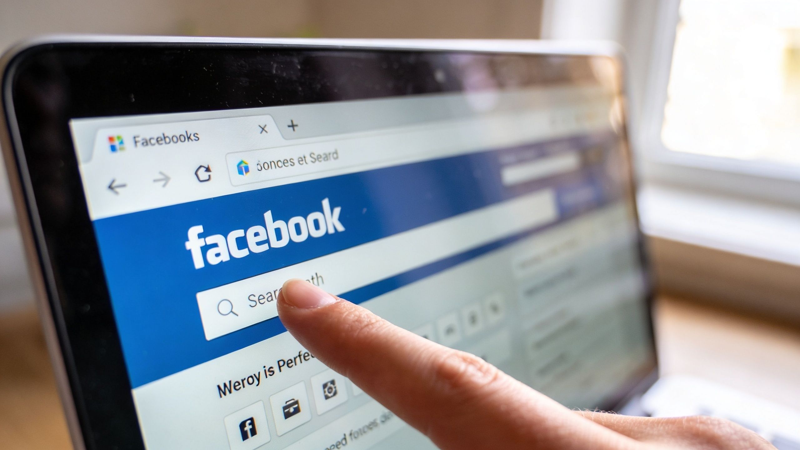 A close-up view of a person using a finger to point at the Facebook search bar on a laptop.