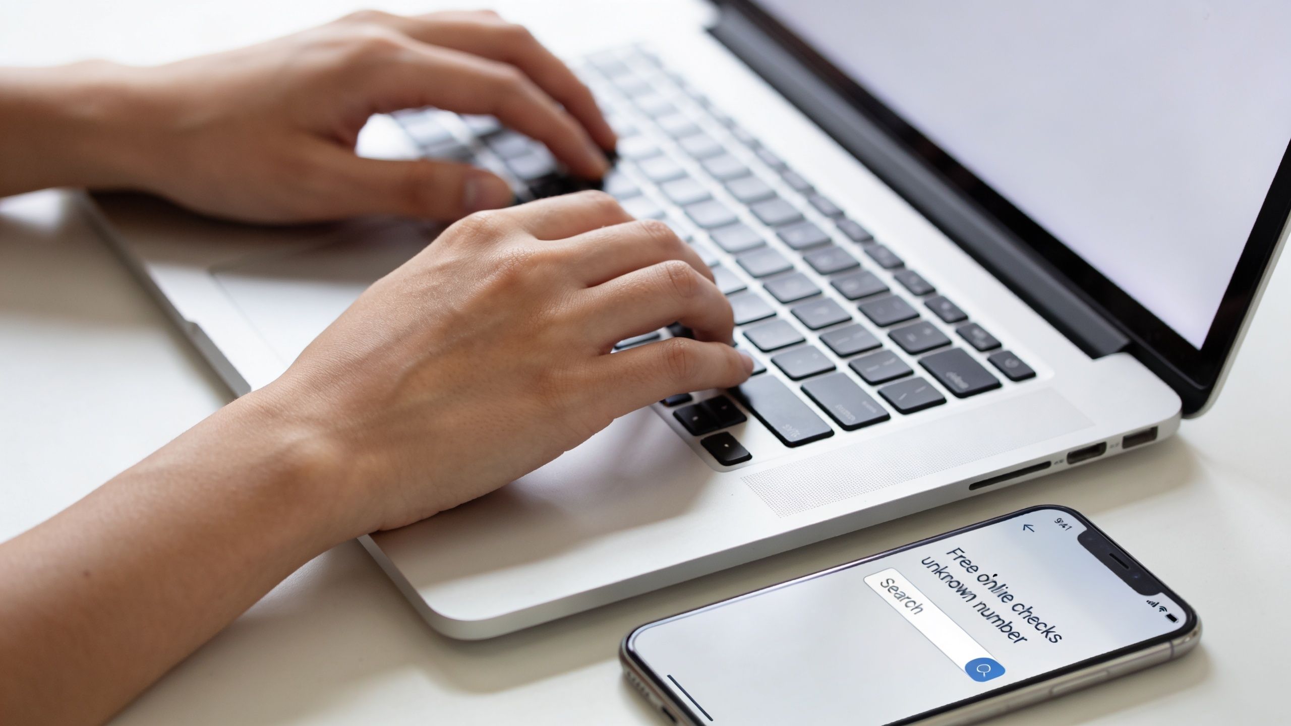 A person using a laptop with a smartphone nearby displaying a search bar for identifying unknown numbers.