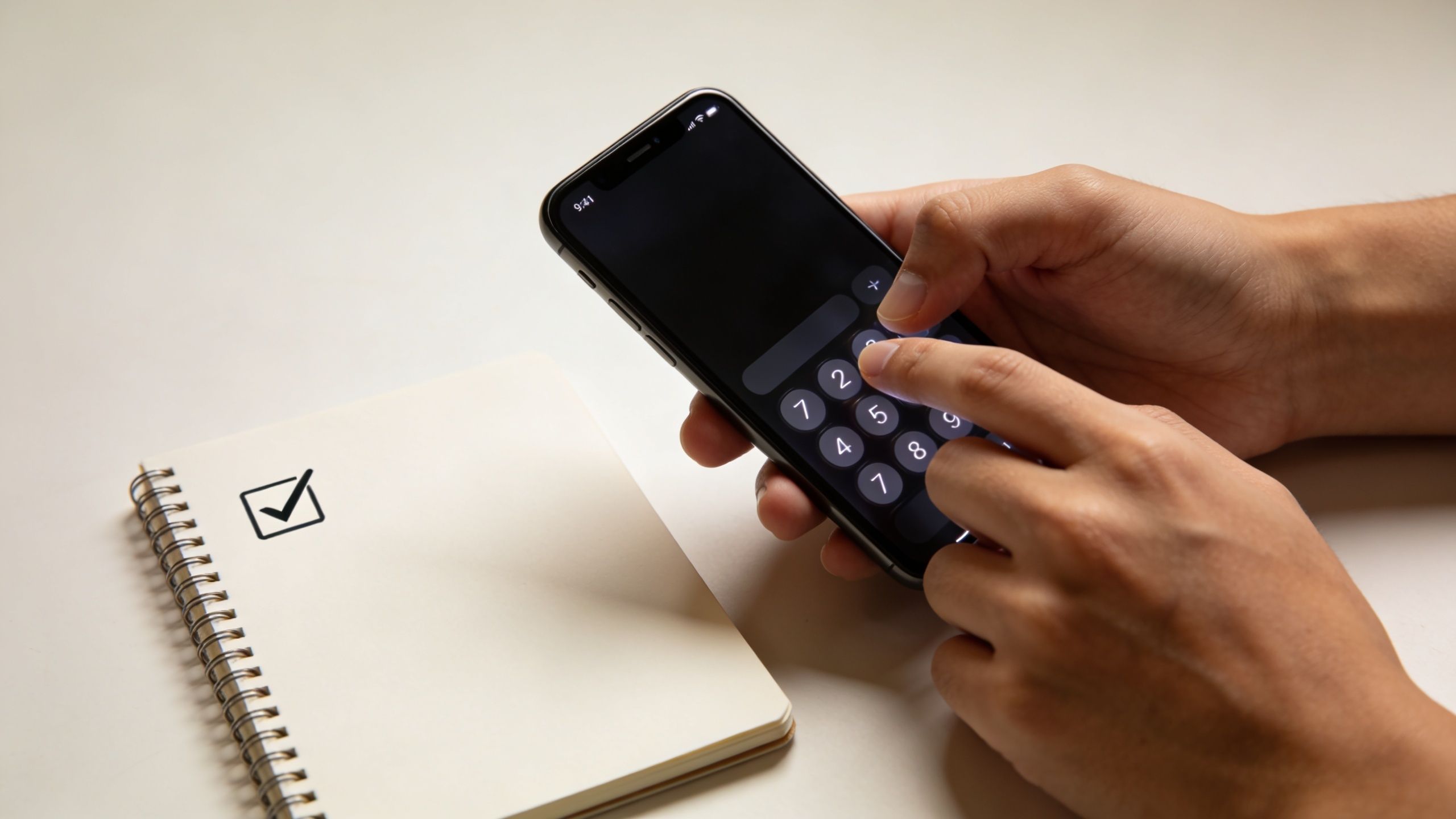A person uses a mobile phone calculator app next to a notebook with a checklist icon.