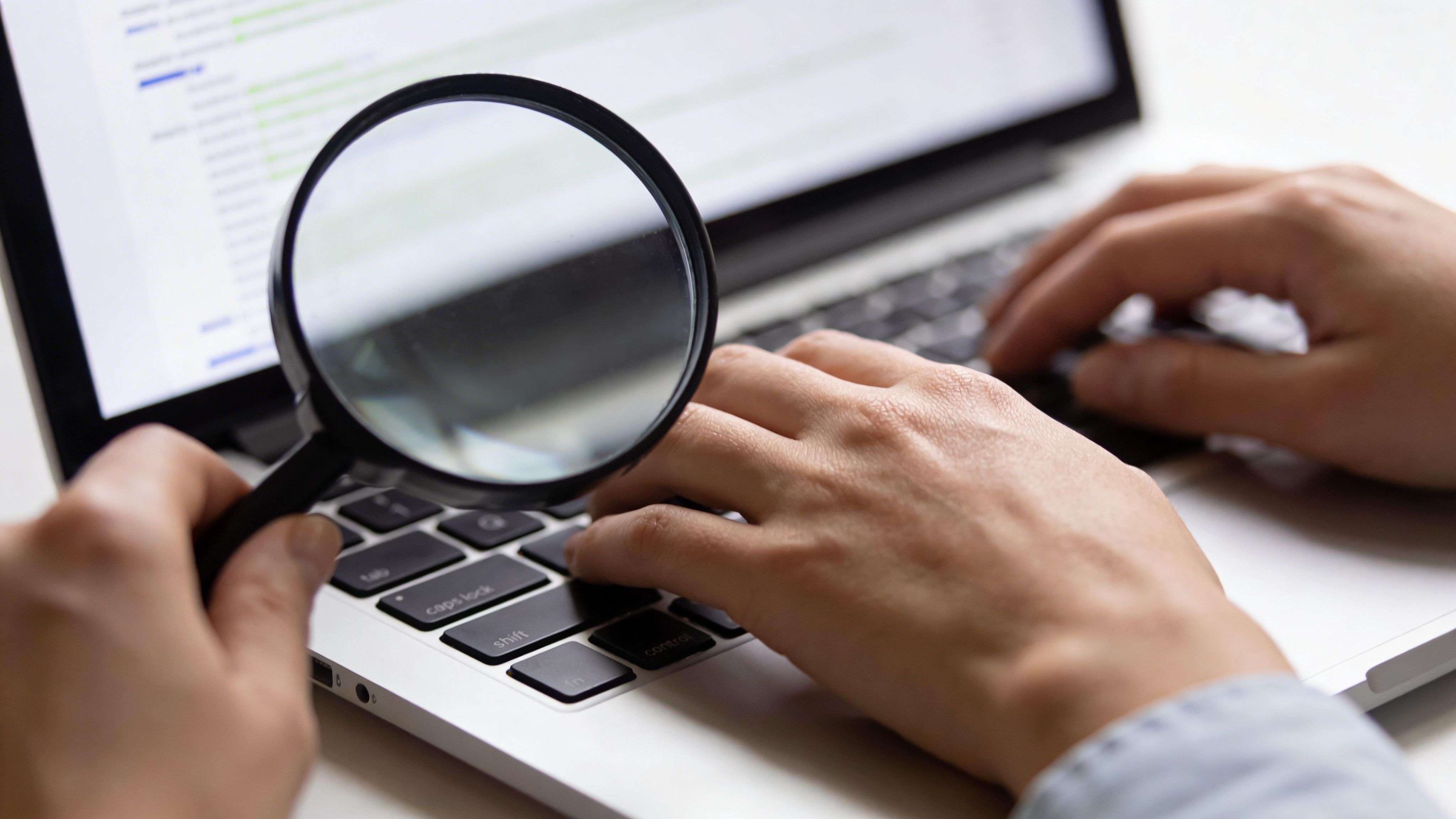 A person uses a magnifying glass to inspect data on a laptop screen while typing on the keyboard.