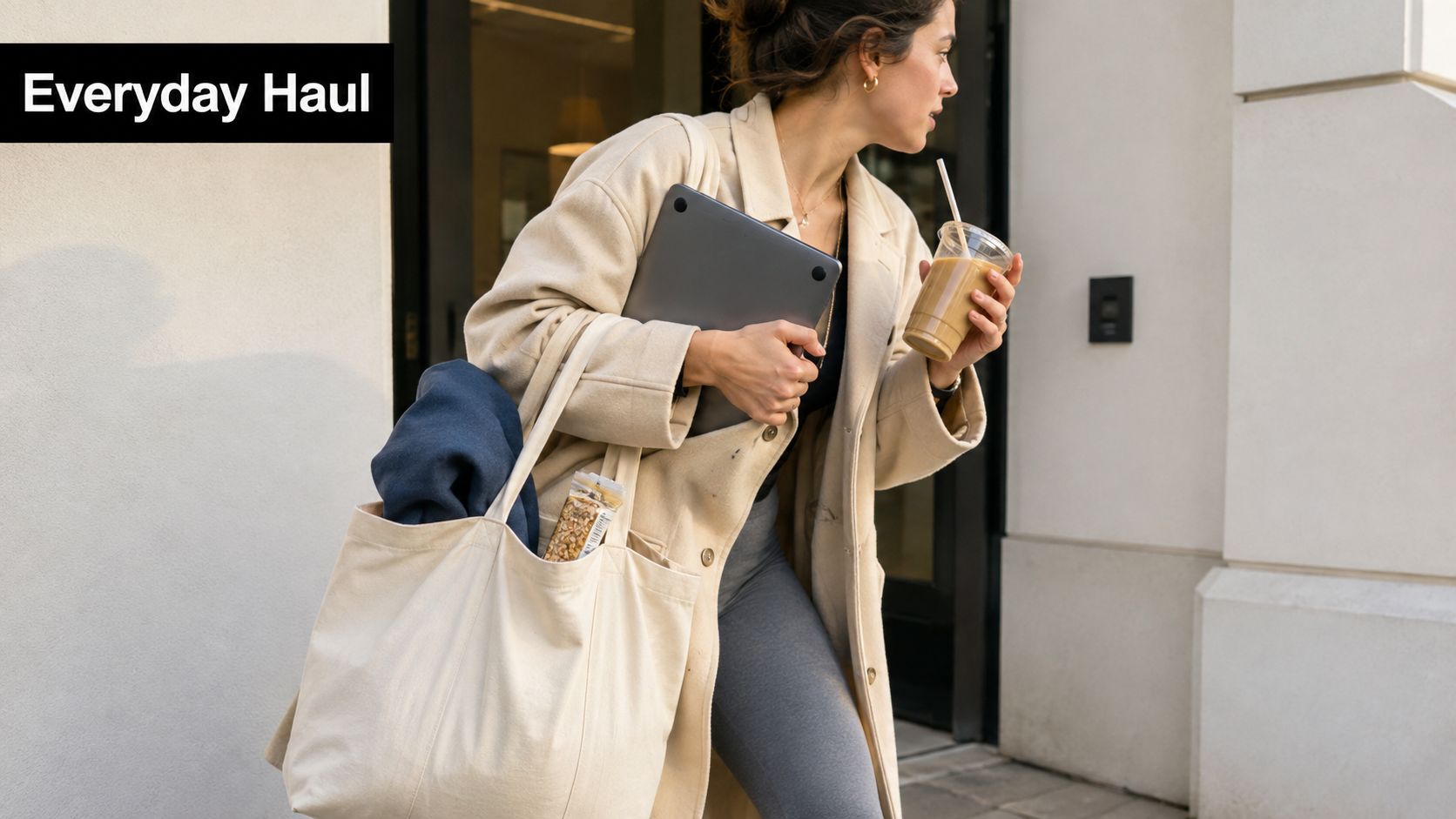 A woman carrying a canvas tote bag, a laptop, and an iced coffee walking through a doorway.