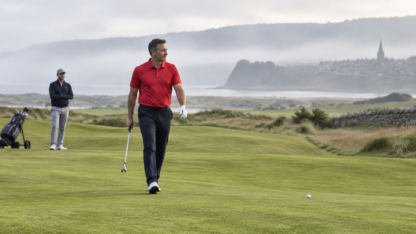 A golfer in a red shirt walks across a scenic golf course towards his ball.
