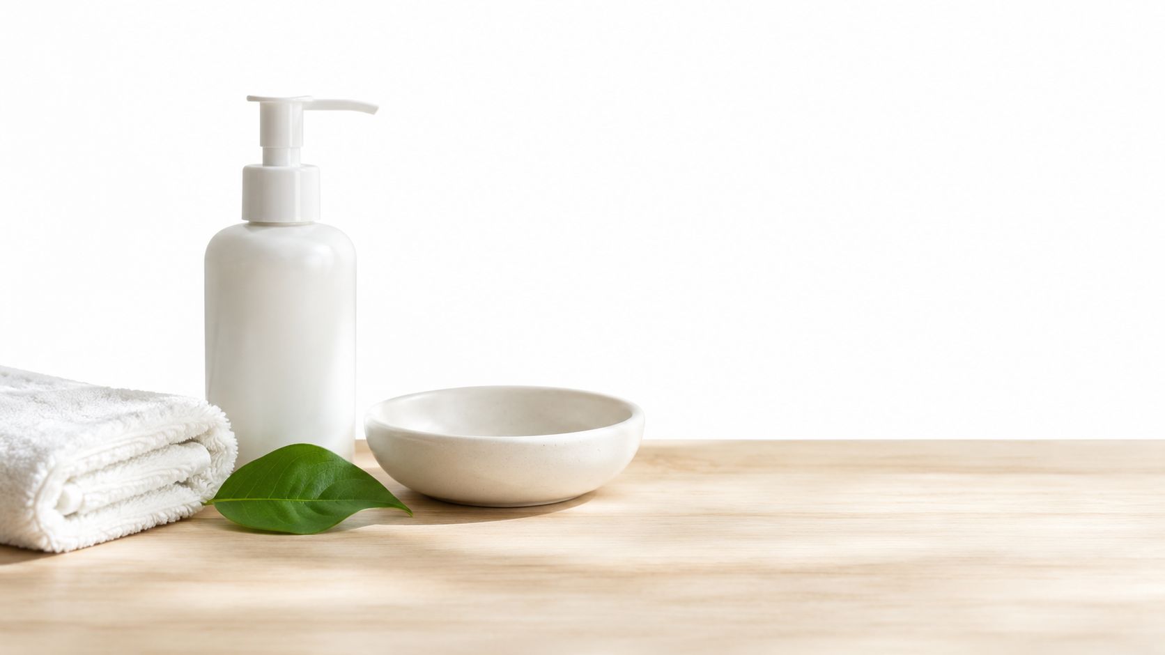 A white pump bottle, a ceramic bowl, and a towel on a wooden surface with a leaf.