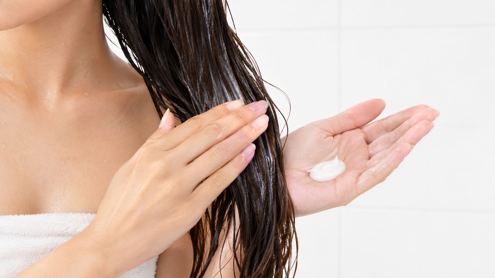 A woman applying hair conditioner to her wet hair in a bathroom setting.
