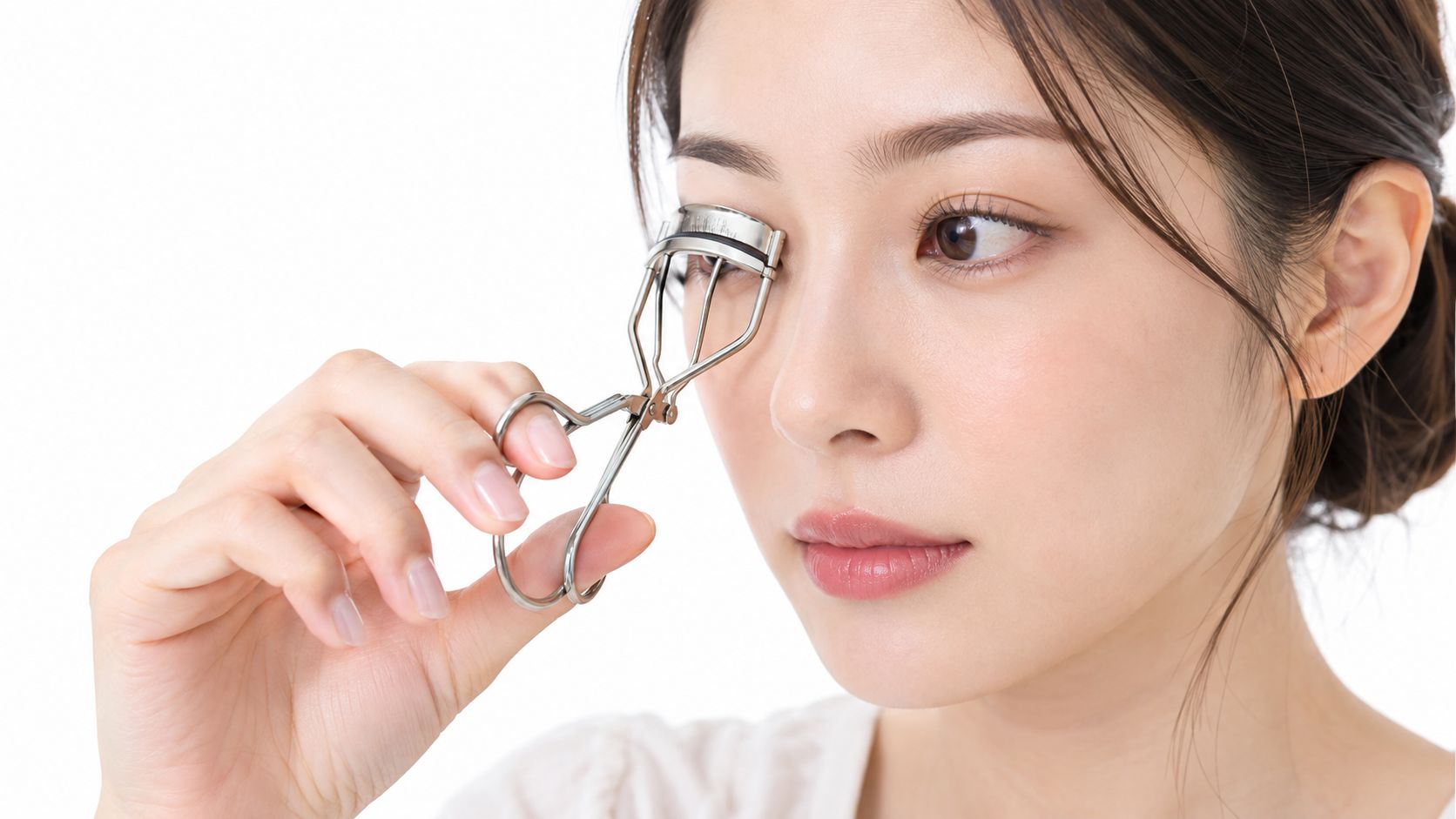 A young Asian woman carefully using a metal eyelash curler to style her eyelashes before applying mascara.