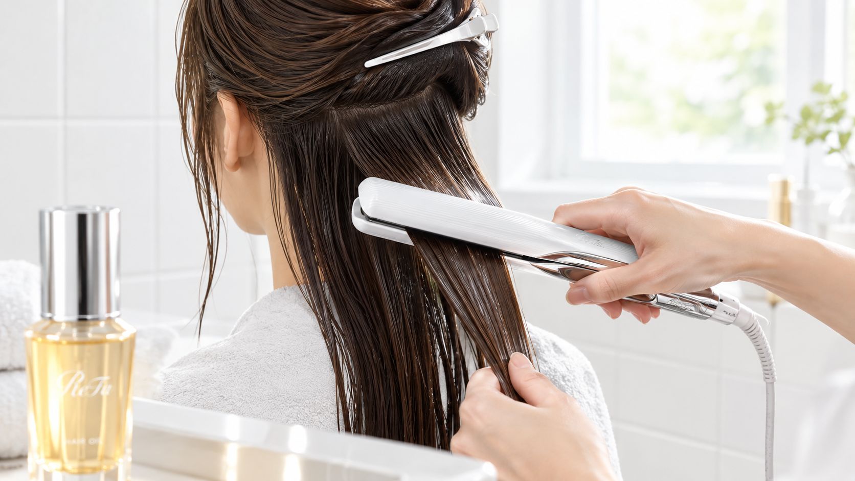 A person using a ReFa flat iron to style damp hair with a bottle of ReFa oil nearby.