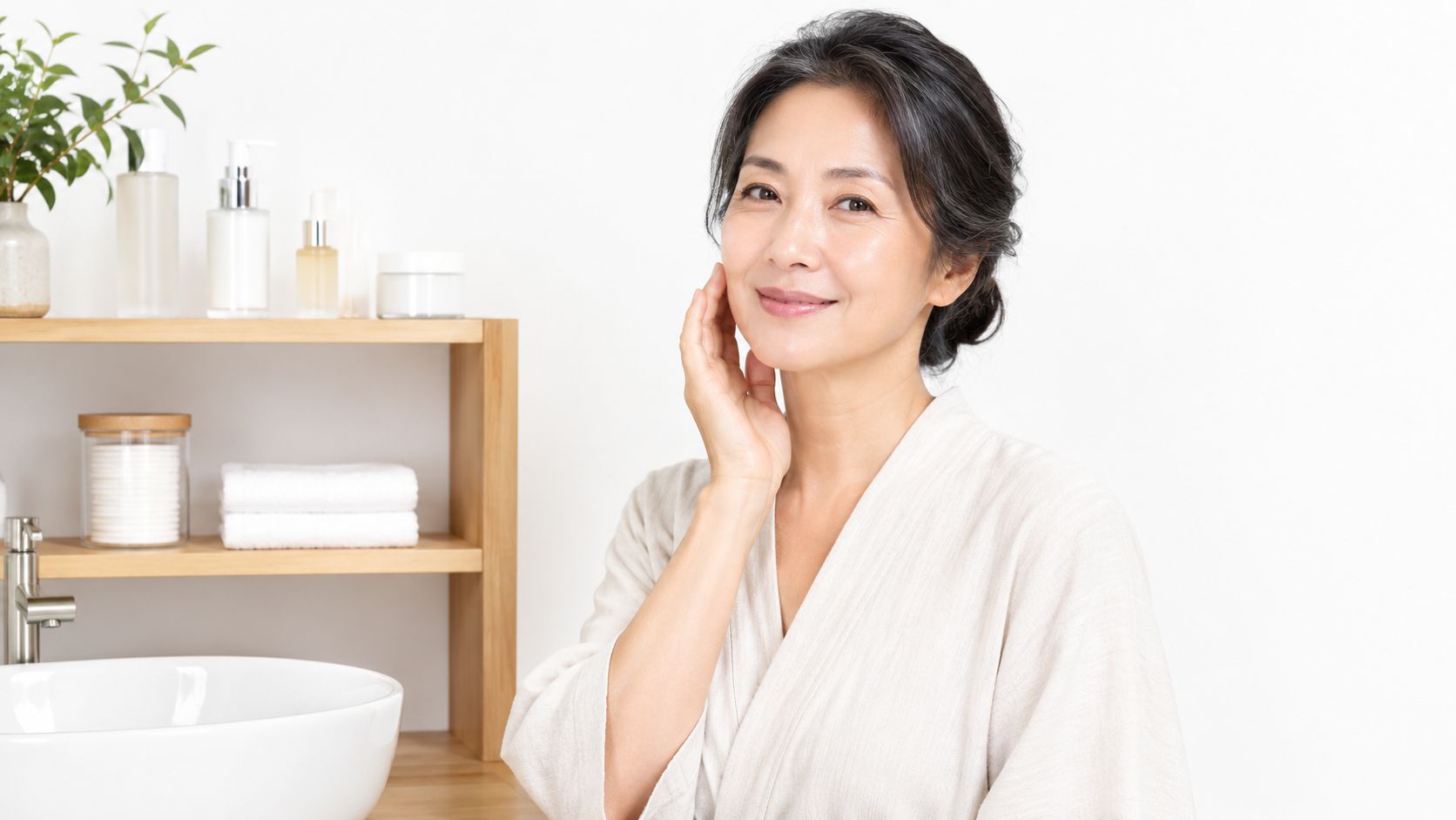 A middle-aged Japanese woman with glowing skin smiling while touching her face in a bright bathroom.