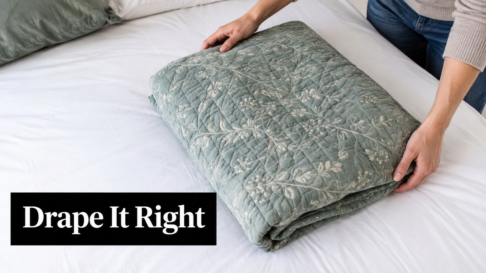 A person carefully folding a soft, sage green floral quilt on a white bed sheet.