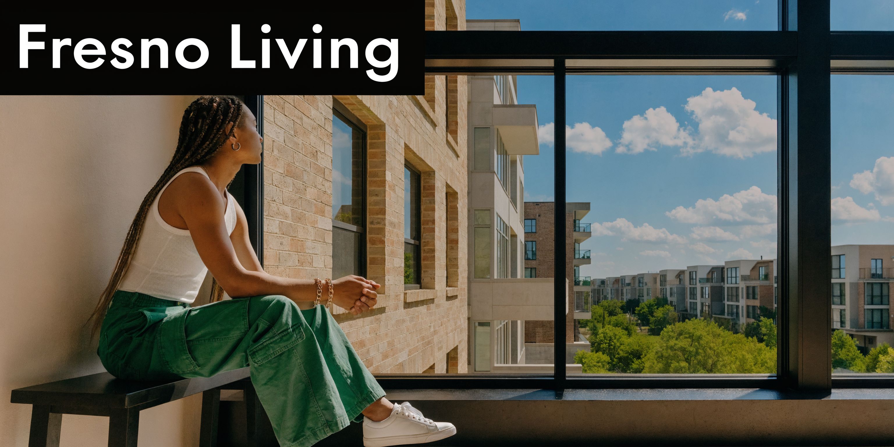 A young woman with long braids sitting on a window bench looking out at modern townhouses.