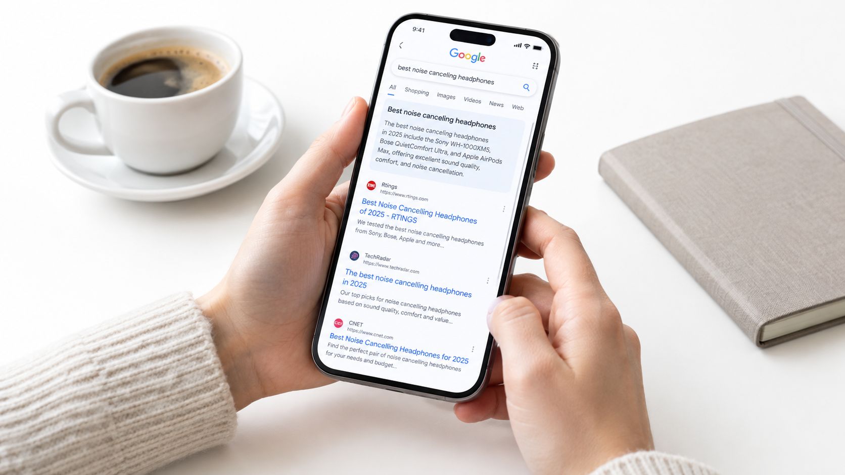 A person holding a smartphone showing Google search results for best noise canceling headphones on a desk.