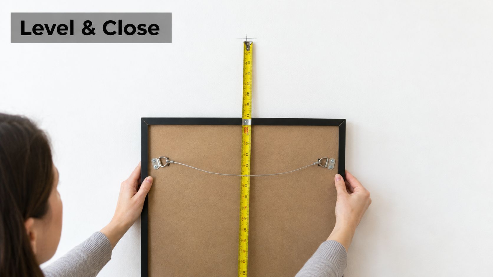 A person holding a framed picture against a white wall while using a measuring tape for alignment.