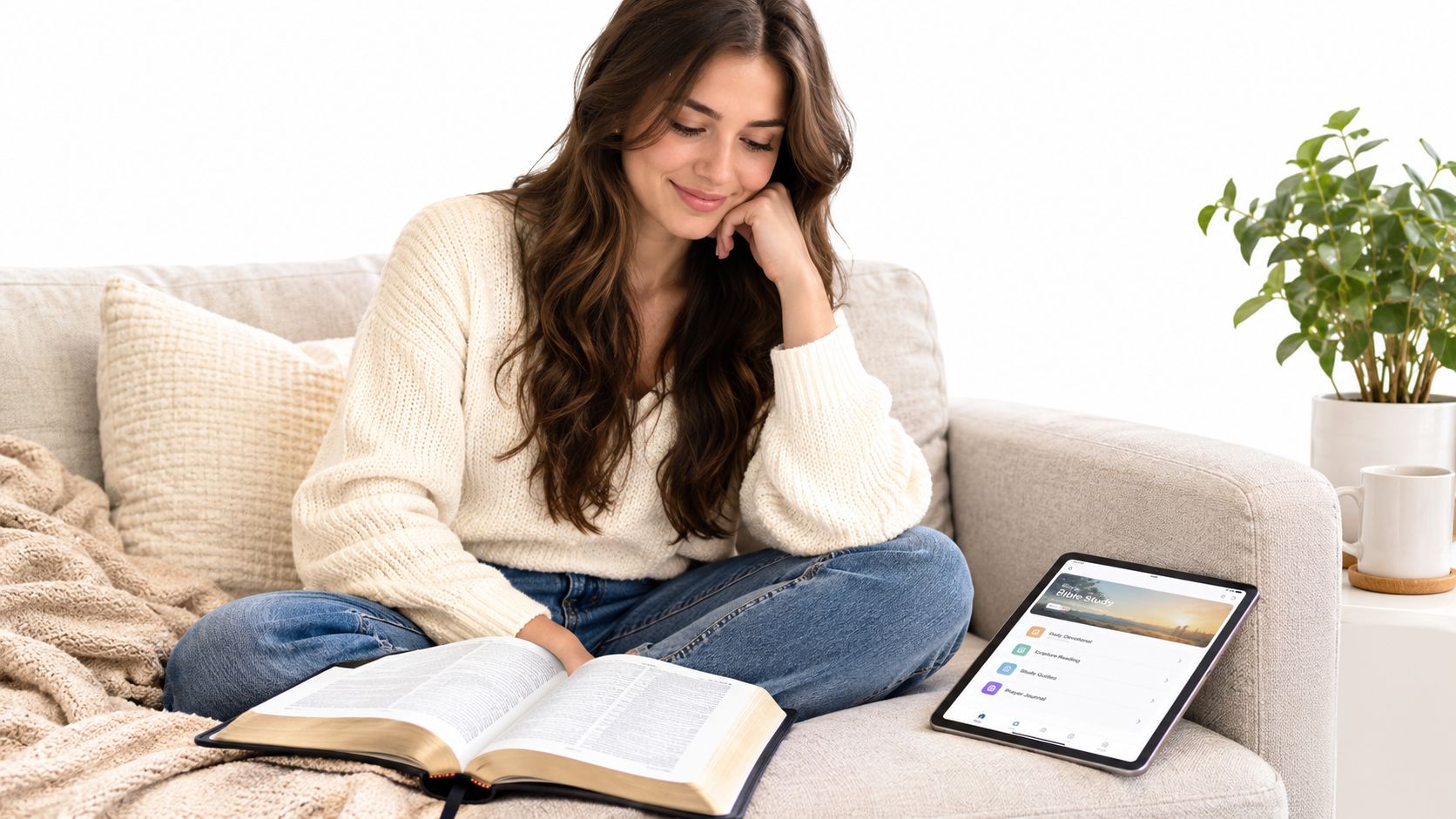 A young woman sitting on a cozy couch reading a physical Bible alongside a digital tablet.