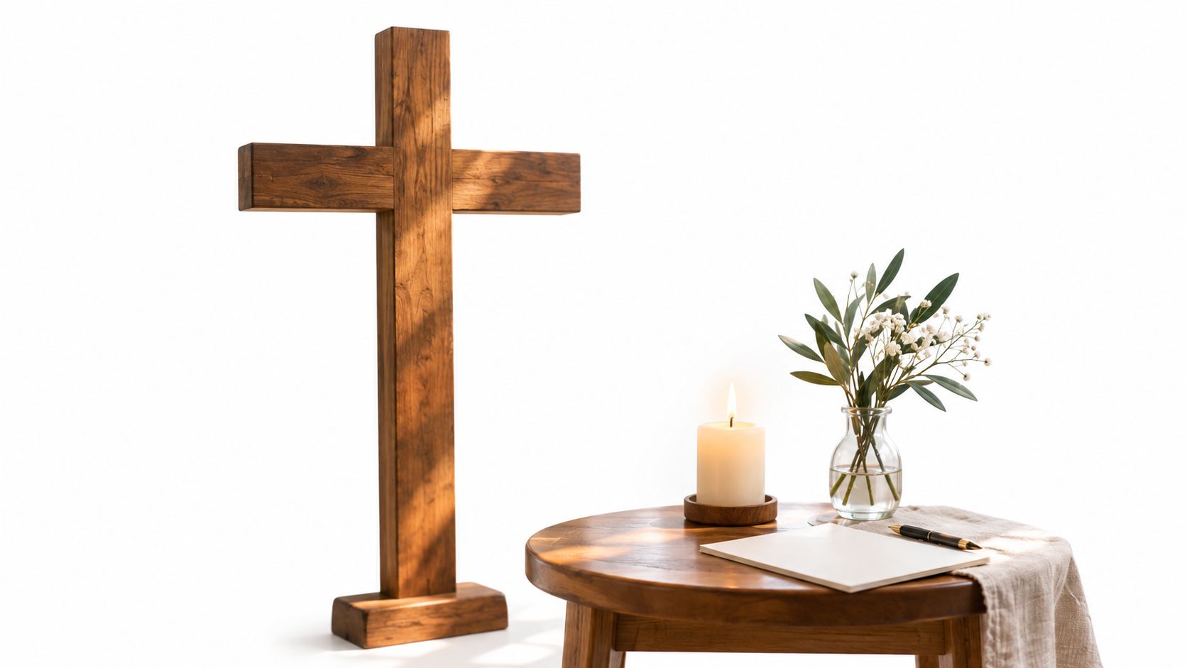 A wooden cross, a lit candle, and a vase with greenery arranged on a small wooden table.
