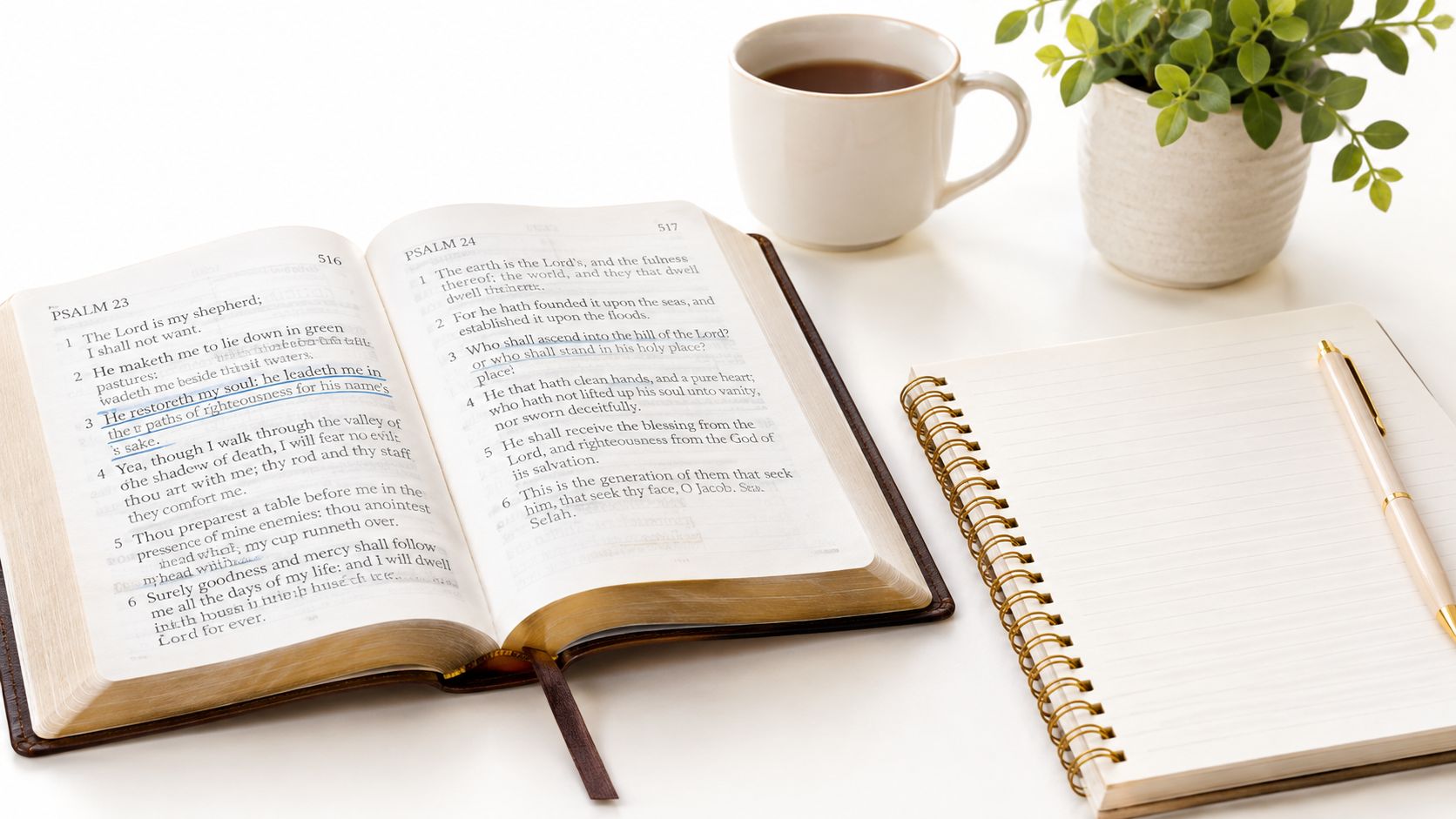 An open Bible showing Psalm 23 and 24 next to a blank notebook, coffee, and a plant.