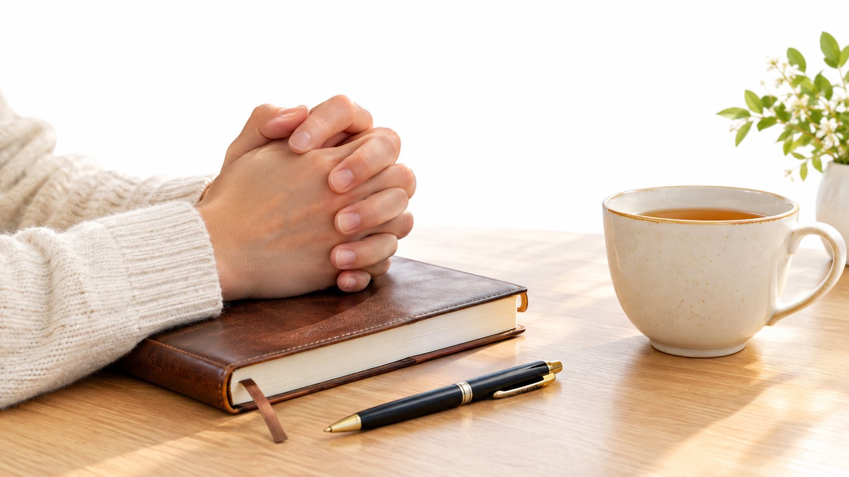 A person with clasped hands resting on a closed leather-bound Bible beside a pen and tea.