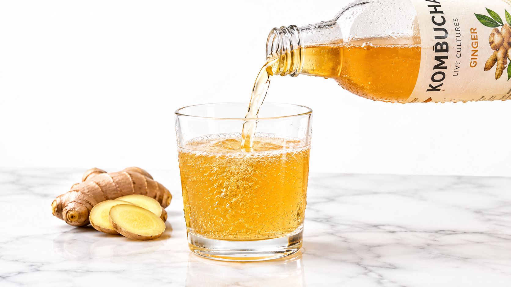 A glass of sparkling ginger kombucha being poured from a bottle beside fresh ginger root slices.