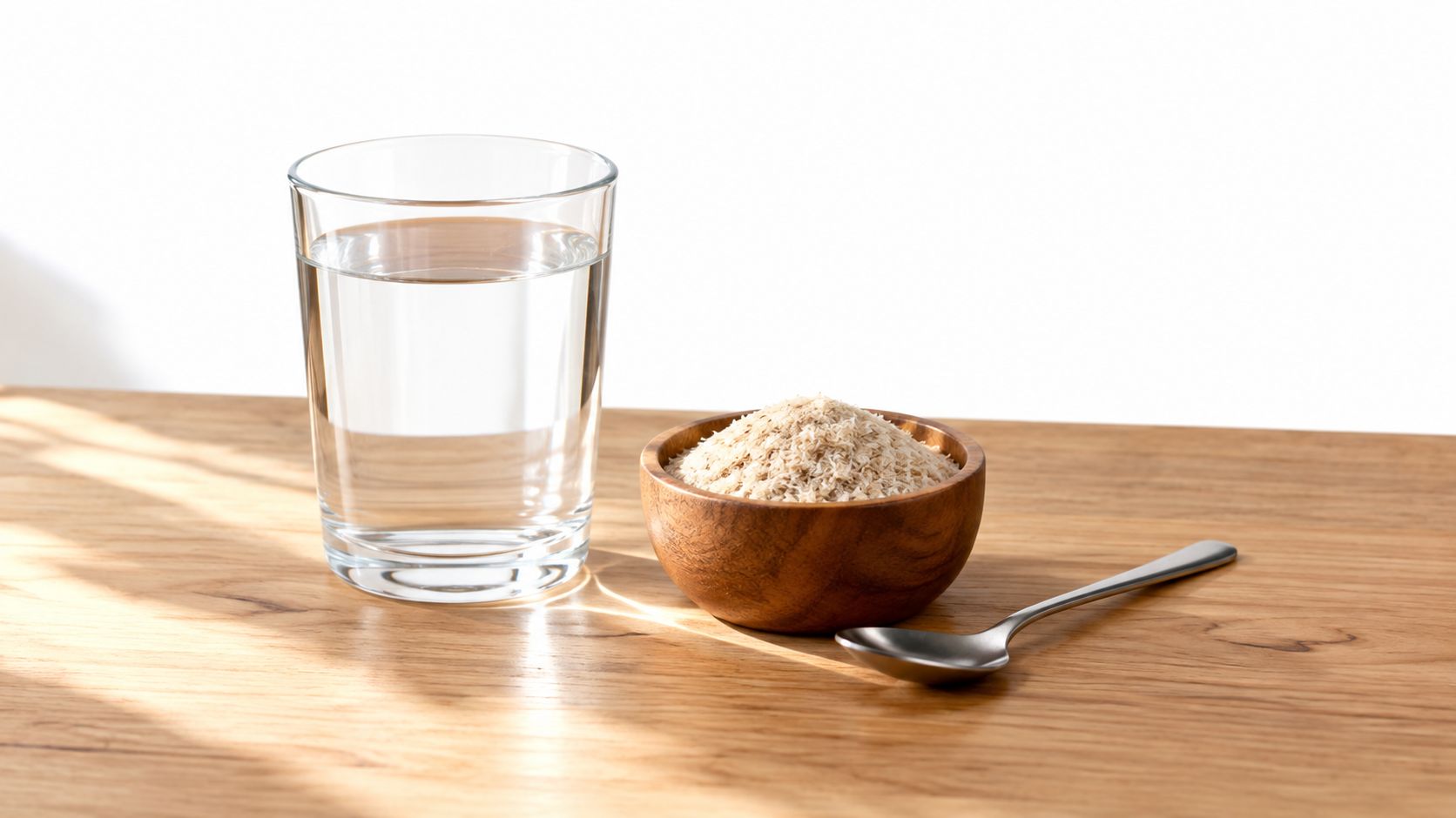 A glass of water next to a wooden bowl containing psyllium husks and a silver spoon.
