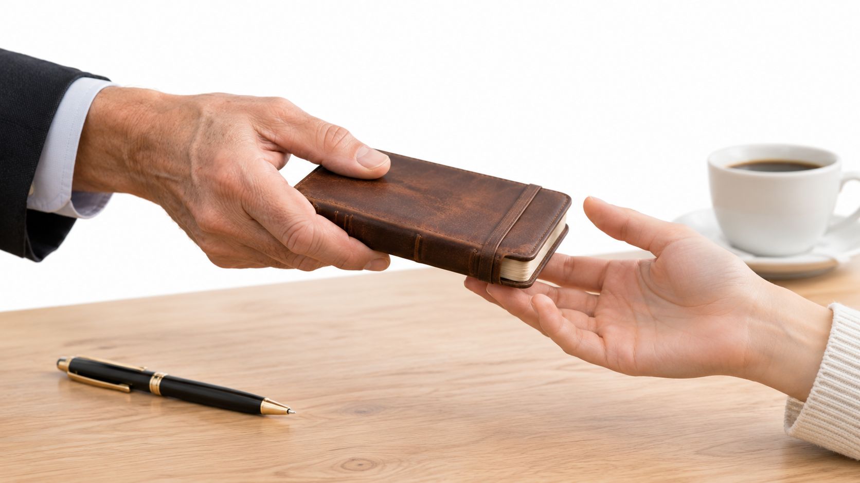 A professional hand passing a small leather journal to another person across a wooden office desk.