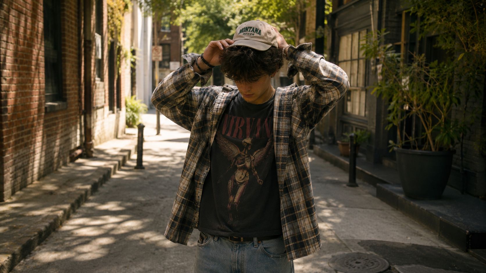 A young man wearing a Nirvana t-shirt, flannel shirt, and a beige Montana dad hat outdoors.