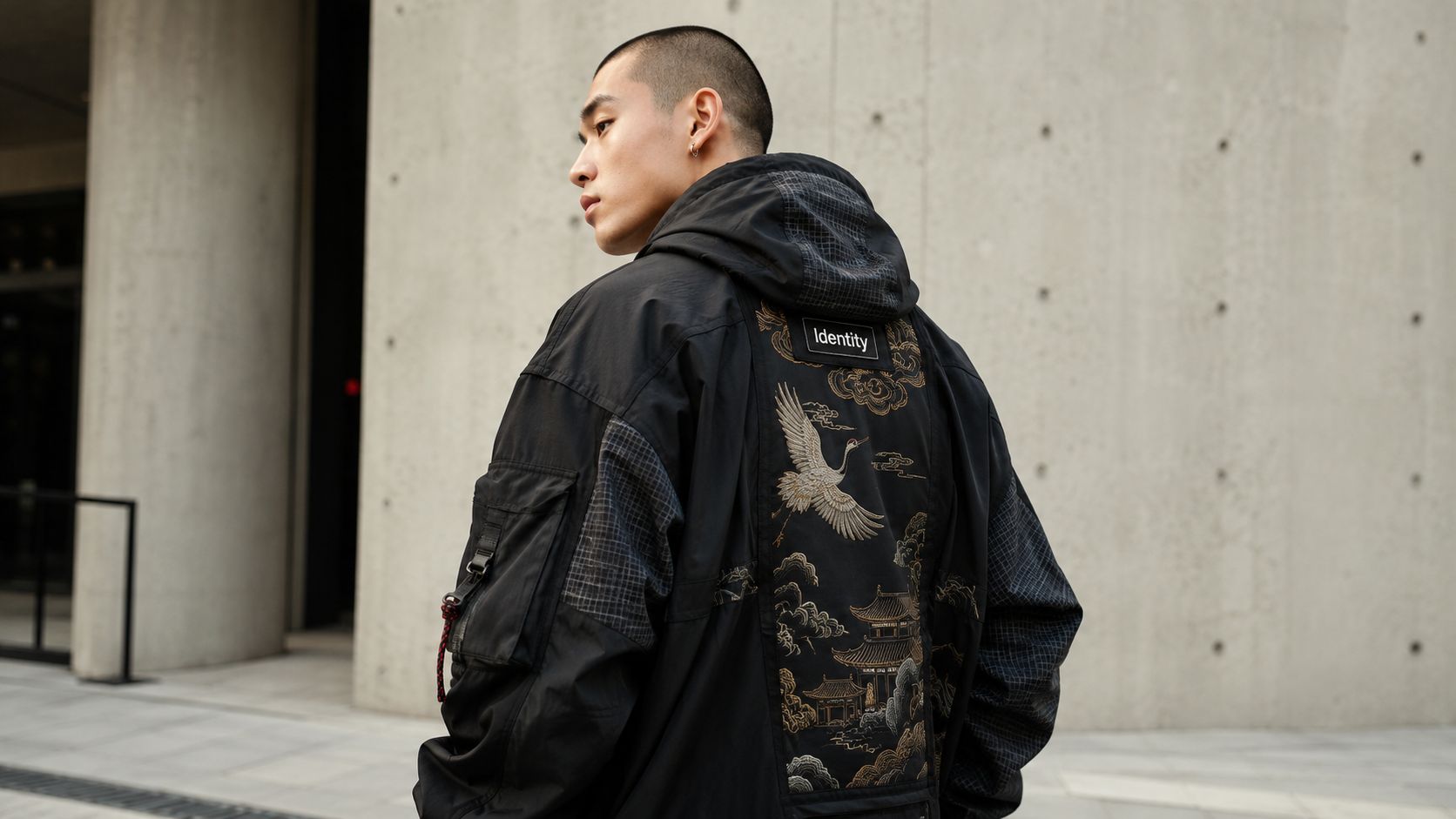 A young man posing in a black techwear jacket featuring traditional crane and temple embroidery.