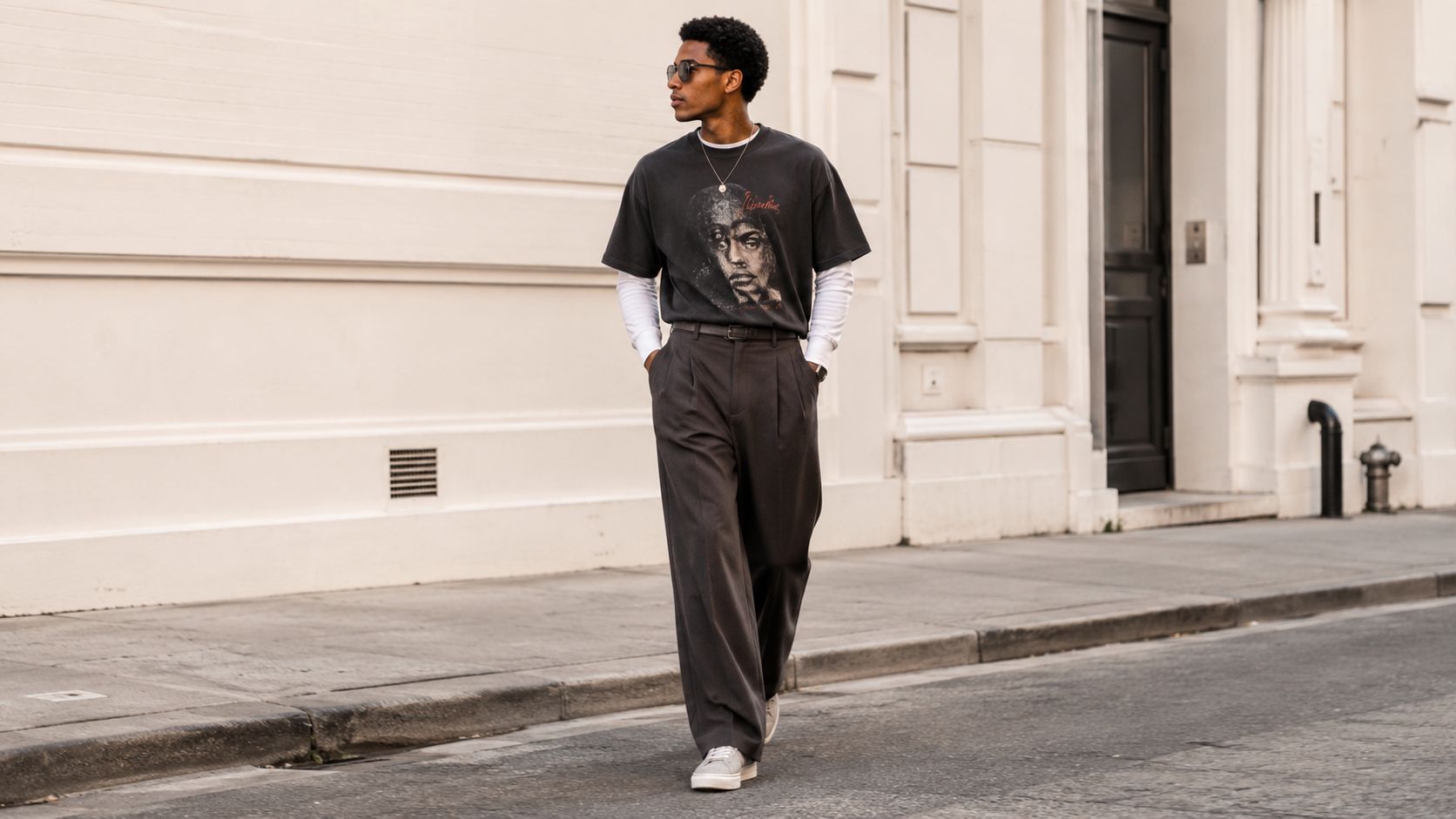A stylish young man wearing an oversized graphic t-shirt and wide-leg trousers walking down a city street.