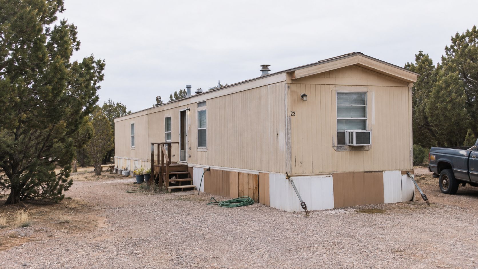 A tan single-wide mobile home with a small wooden porch parked on a gravel lot in nature.