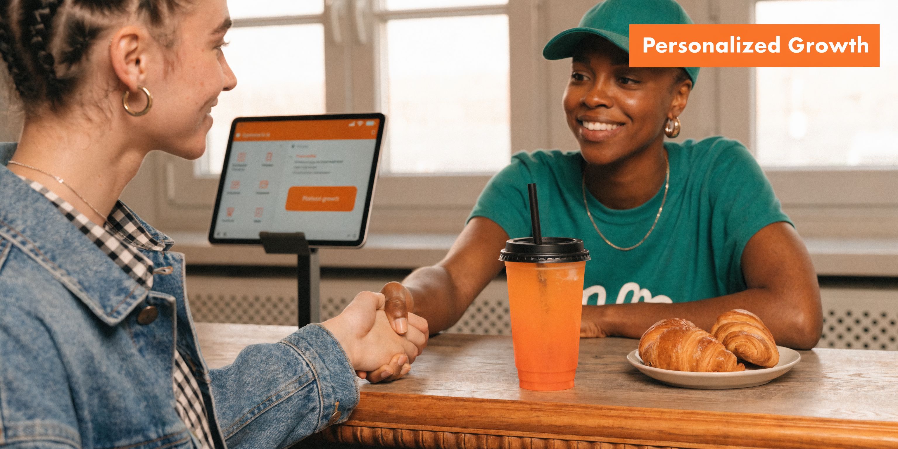 A smiling barista shakes hands with a customer at a cafe counter next to a digital tablet.
